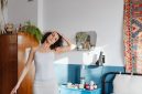 Seorang wanita tersenyum cerah sedang melakukan peregangan ringan di pagi hari, menunjukkan bahwa memulai kebugaran tidak selalu membutuhkan peralatan rumit. Di sebelahnya tampak meja sarapan sehat penuh buah segar dan minuman, melambangkan pentingnya nutrisi seimbang untuk energi sepanjang hari.