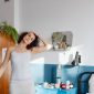 Seorang wanita tersenyum cerah sedang melakukan peregangan ringan di pagi hari, menunjukkan bahwa memulai kebugaran tidak selalu membutuhkan peralatan rumit. Di sebelahnya tampak meja sarapan sehat penuh buah segar dan minuman, melambangkan pentingnya nutrisi seimbang untuk energi sepanjang hari.
