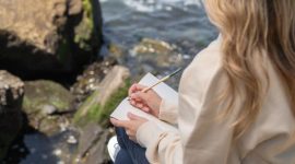 Seorang wanita tersenyum damai sambil menulis di jurnal, mencerminkan momen refleksi diri dan penemuan kebahagiaan di tengah alam yang menenangkan. Kegiatan menulis jurnal dapat menjadi salah satu ritual penting untuk memupuk kebahagiaan dan mengidentifikasi nilai-nilai hidup yang sesungguhnya.