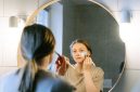 Seorang wanita muda Asia sedang mengaplikasikan produk skincare pada wajahnya di kamar mandi yang terang, menunjukkan fokus pada rutinitas perawatan diri untuk mendapatkan kulit sehat dan bercahaya. Foto ini merefleksikan tren kecantikan Korea yang mengedepankan hidrasi dan langkah perawatan yang teratur.