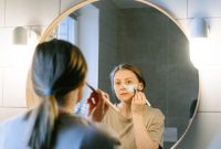 Seorang wanita muda Asia sedang mengaplikasikan produk skincare pada wajahnya di kamar mandi yang terang, menunjukkan fokus pada rutinitas perawatan diri untuk mendapatkan kulit sehat dan bercahaya. Foto ini merefleksikan tren kecantikan Korea yang mengedepankan hidrasi dan langkah perawatan yang teratur.