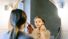 Seorang wanita muda Asia sedang mengaplikasikan produk skincare pada wajahnya di kamar mandi yang terang, menunjukkan fokus pada rutinitas perawatan diri untuk mendapatkan kulit sehat dan bercahaya. Foto ini merefleksikan tren kecantikan Korea yang mengedepankan hidrasi dan langkah perawatan yang teratur.
