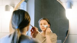 Seorang wanita muda Asia sedang mengaplikasikan produk skincare pada wajahnya di kamar mandi yang terang, menunjukkan fokus pada rutinitas perawatan diri untuk mendapatkan kulit sehat dan bercahaya. Foto ini merefleksikan tren kecantikan Korea yang mengedepankan hidrasi dan langkah perawatan yang teratur.