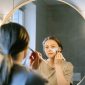 Seorang wanita muda Asia sedang mengaplikasikan produk skincare pada wajahnya di kamar mandi yang terang, menunjukkan fokus pada rutinitas perawatan diri untuk mendapatkan kulit sehat dan bercahaya. Foto ini merefleksikan tren kecantikan Korea yang mengedepankan hidrasi dan langkah perawatan yang teratur.