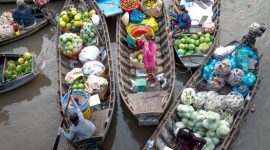 Pemandangan ramai aktivitas jual beli di pasar terapung, menampilkan perahu-perahu kecil penuh dengan aneka dagangan hasil bumi dan kerajinan tangan, berinteraksi langsung dengan pembeli di atas sungai. Suasana pagi yang cerah menambah keindahan interaksi budaya yang telah berlangsung turun-temurun ini.