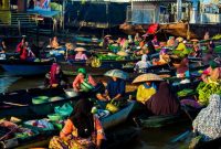 Deretan perahu jukung berisi beragam dagangan segar mengapung di sungai, menyajikan pemandangan khas pasar terapung di Kalimantan yang ramai dengan aktivitas jual beli. Para pedagang dengan cekatan melayani pembeli, menciptakan suasana interaksi yang hidup dan penuh warna di atas air.