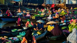 Deretan perahu jukung berisi beragam dagangan segar mengapung di sungai, menyajikan pemandangan khas pasar terapung di Kalimantan yang ramai dengan aktivitas jual beli. Para pedagang dengan cekatan melayani pembeli, menciptakan suasana interaksi yang hidup dan penuh warna di atas air.