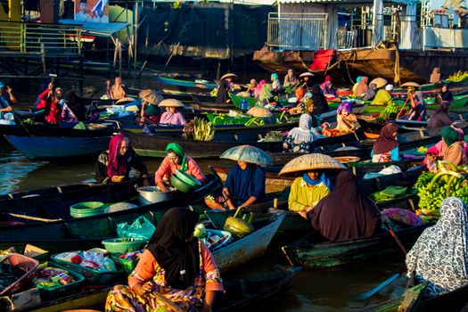 Deretan perahu jukung berisi beragam dagangan segar mengapung di sungai, menyajikan pemandangan khas pasar terapung di Kalimantan yang ramai dengan aktivitas jual beli. Para pedagang dengan cekatan melayani pembeli, menciptakan suasana interaksi yang hidup dan penuh warna di atas air.