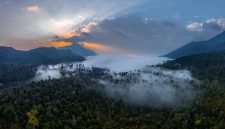Pemandangan rimbun Pegunungan Meratus yang diselimuti kabut, memperlihatkan rumah adat suku Dayak Meratus yang menyatu harmonis dengan lingkungan, menggambarkan kehidupan tradisional yang masih terjaga.
