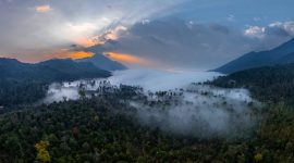 Pemandangan rimbun Pegunungan Meratus yang diselimuti kabut, memperlihatkan rumah adat suku Dayak Meratus yang menyatu harmonis dengan lingkungan, menggambarkan kehidupan tradisional yang masih terjaga.