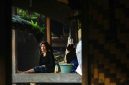 Seorang warga Dayak Meratus berdiri di depan rumah adat mereka yang kokoh, melambangkan kuatnya akar tradisi dan kebersamaan dalam menghadapi tantangan zaman. Bangunan ini tidak hanya tempat tinggal, tetapi juga pusat aktivitas sosial dan spiritual komunitas.